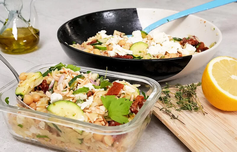 Im Vordergrund steht in einer Glasbox Orzo-Salat mit Zucchini und Kichererbsen mit Honig-Senf-Dressing nach einem Rezept von Felicitas Then. Im Hintergrund ist der Orzo-Salat auf einem schwarz-weißen Teller angerichtet. Rechts liegt eine halbe Zitrone, hinten links steht ein Ölkännchen.