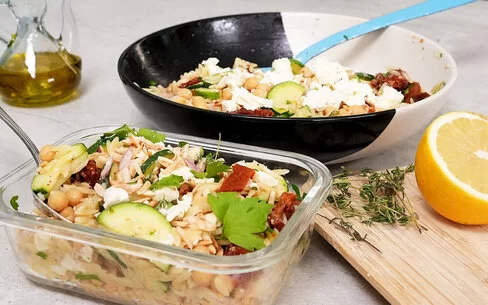 Im Vordergrund steht in einer Glasbox Orzo-Salat mit Zucchini und Kichererbsen mit Honig-Senf-Dressing nach einem Rezept von Felicitas Then. Im Hintergrund ist der Orzo-Salat auf einem schwarz-weißen Teller angerichtet. Rechts liegt eine halbe Zitrone, hinten links steht ein Ölkännchen.