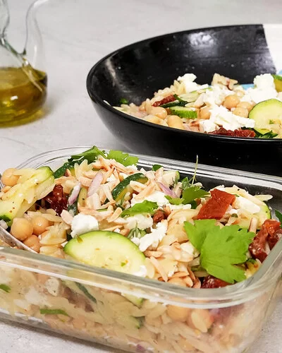 Im Vordergrund steht in einer Glasbox Orzo-Salat mit Zucchini und Kichererbsen mit Honig-Senf-Dressing nach einem Rezept von Felicitas Then. Im Hintergrund ist der Orzo-Salat auf einem schwarz-weißen Teller angerichtet. Rechts liegt eine halbe Zitrone, hinten links steht ein Ölkännchen.