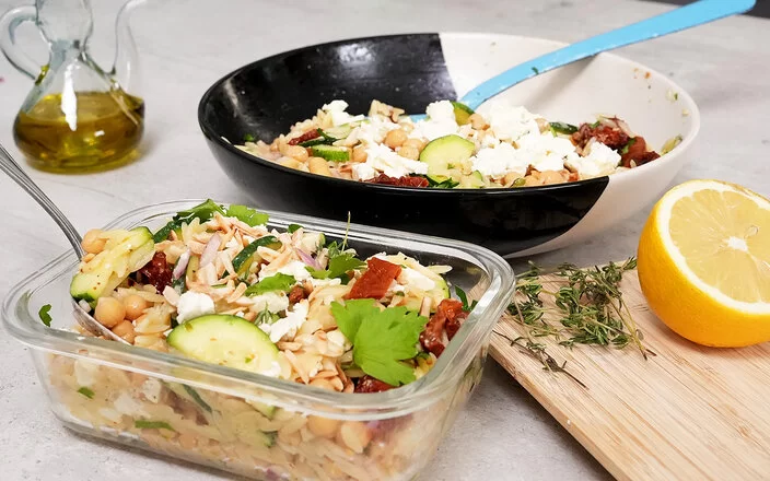 Im Vordergrund steht in einer Glasbox Orzo-Salat mit Zucchini und Kichererbsen mit Honig-Senf-Dressing nach einem Rezept von Felicitas Then. Im Hintergrund ist der Orzo-Salat auf einem schwarz-weißen Teller angerichtet. Rechts liegt eine halbe Zitrone, hinten links steht ein Ölkännchen.