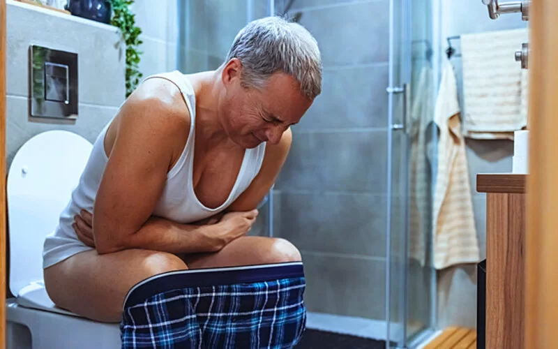 Ein Mann sitzt auf einer Toilette und beugt sich mit geschlossenen Augen leicht nach vorn, während er sich mit den Armen auf den Beinen abstützt.