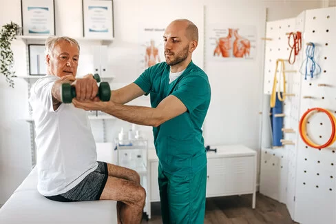 Ein junger Mann stützt den Arm eines älteren Mannes, der gerade eine Hantel hebt. Die AOK unterstützt die Physiotherapie.