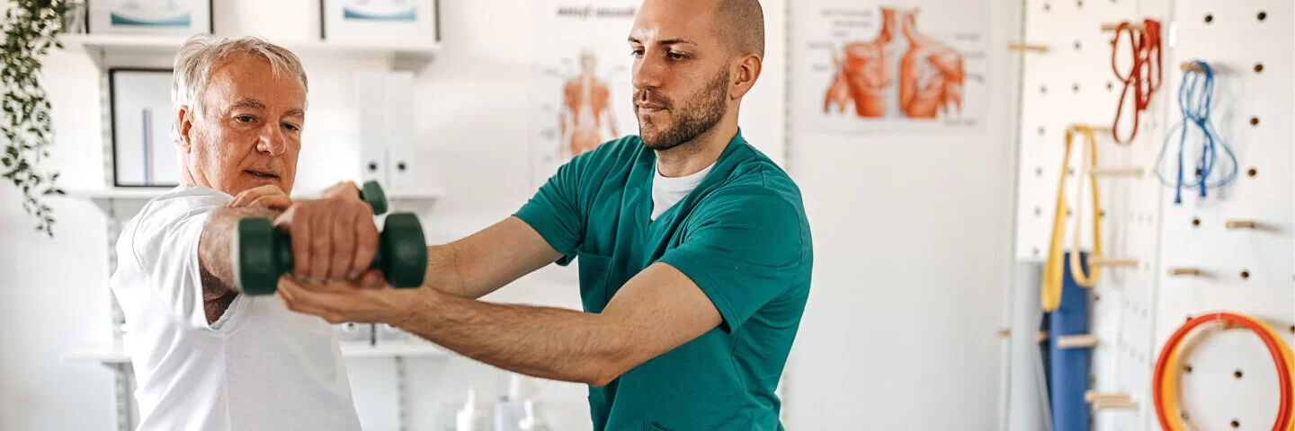 Ein junger Mann stützt den Arm eines älteren Mannes, der gerade eine Hantel hebt. Die AOK unterstützt die Physiotherapie.