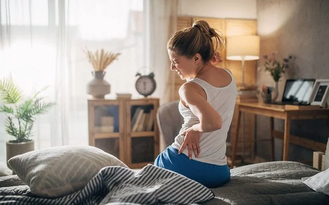 Eine Frau sitzt mit Rückenschmerzen auf der Bettkante und hält sich schmerzverzogen den Rücken fest.