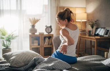 Eine Frau sitzt mit Rückenschmerzen auf der Bettkante und hält sich schmerzverzogen den Rücken fest.