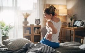 Eine Frau sitzt mit Rückenschmerzen auf der Bettkante und hält sich schmerzverzogen den Rücken fest.