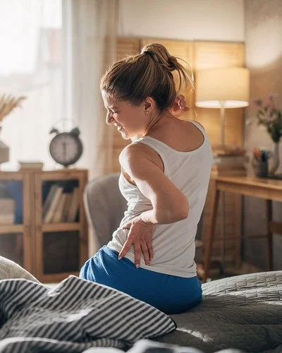 Eine Frau sitzt mit Rückenschmerzen auf der Bettkante und hält sich schmerzverzogen den Rücken fest.