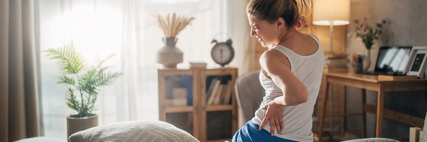 Eine Frau sitzt mit Rückenschmerzen auf der Bettkante und hält sich schmerzverzogen den Rücken fest.