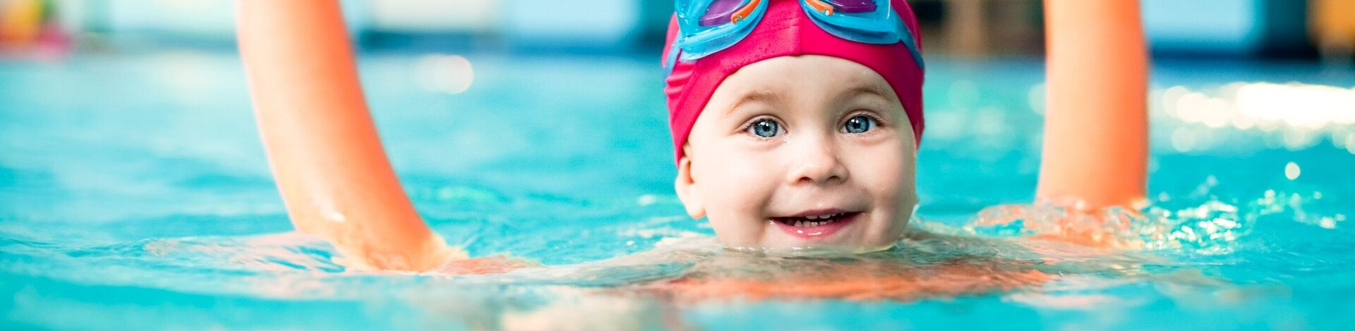 Kleinkind mit Poolnudel und Badekappe schwimmt im Wasser und freut sich.