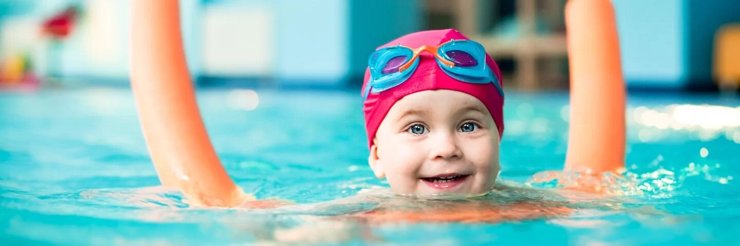 Kleinkind mit Poolnudel und Badekappe schwimmt im Wasser und freut sich.