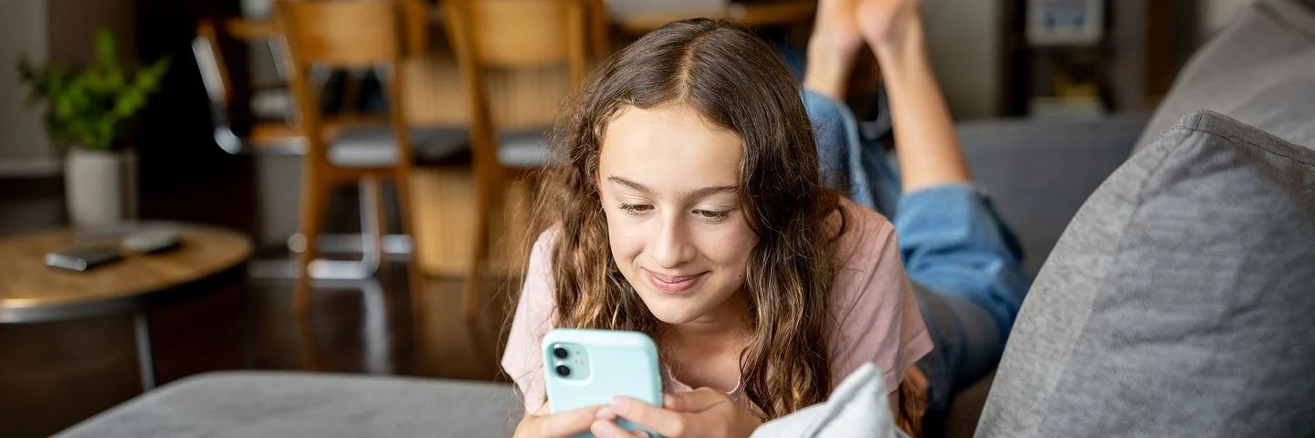 Ein dunkelhaariges Mädchen liegt auf dem Sofa und schaut lächelnd auf sein Smartphone.