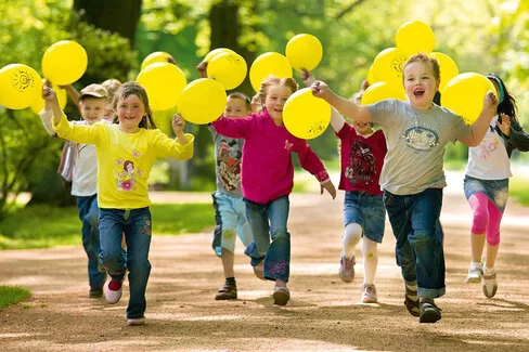 Lachende Kinder rennen bei Sonnenschein mit gelben Luftballons in der Hand durch einen Wald.