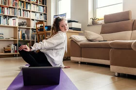 Ein junges Mädchen sitzt auf dem Boden auf ener Matte, schaut in den Bildschirm eines Laptops und macht dabei Yogaübungen.