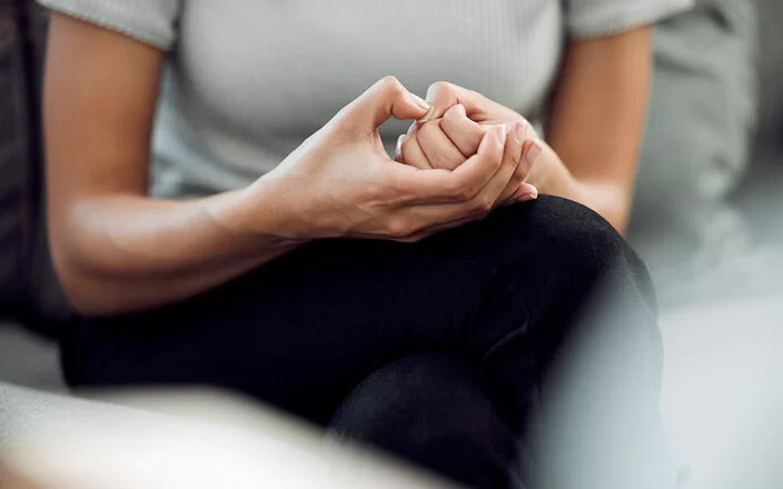 Eine nicht erkennbare junge Frau mit Skin-Picking-Disorder sitzt mit überschlagenen Beinen auf einem Sofa und knibbelt nervös an ihrem Daumennagel.