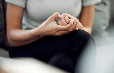 Eine nicht erkennbare junge Frau mit Skin-Picking-Disorder sitzt mit überschlagenen Beinen auf einem Sofa und knibbelt nervös an ihrem Daumennagel.
