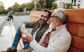 Zwei Männer sitzen entspannt auf einer Holzbank in der Abendsonne und stoßen mit zwei Gläsern Bier an.
