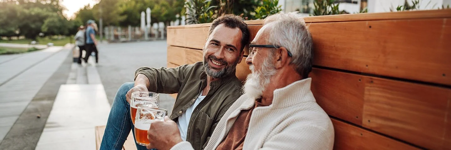 Zwei Männer sitzen entspannt auf einer Holzbank in der Abendsonne und stoßen mit zwei Gläsern Bier an.