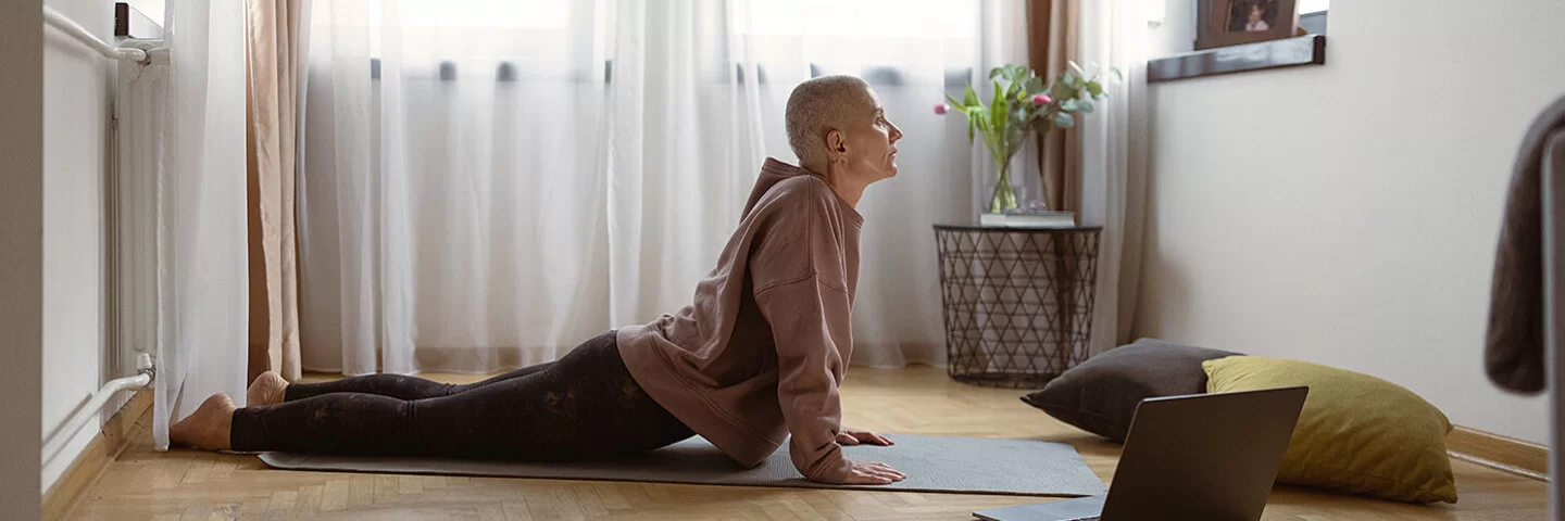 Eine mittelalte Frau mit rasiertem Kopf macht in einem Zimmer auf einer Matte eine Yoga-Stellung, rechts von ihr steht ein geöffnetes Notebook auf dem Boden.