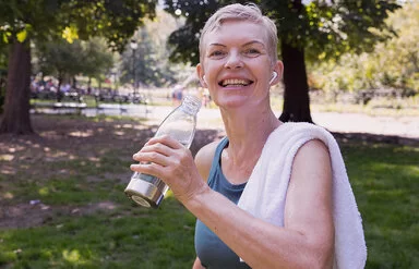 Eine mittelalte Frau mit kurzen blonden Haaren steht nach dem Training lachendlächelnd im Park mit einem über die Schulter geworfenem Handtuch und einer Wasserflasche in der Hand.
