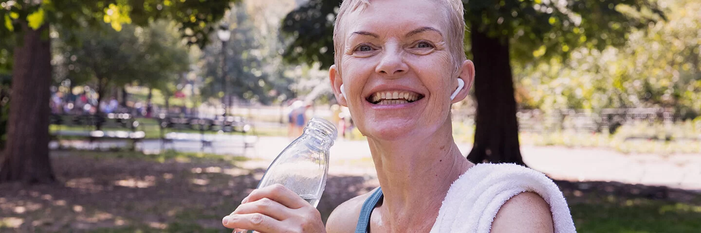 Eine mittelalte Frau mit kurzen blonden Haaren steht nach dem Training lachendlächelnd im Park mit einem über die Schulter geworfenem Handtuch und einer Wasserflasche in der Hand.