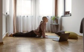 Eine mittelalte Frau mit rasiertem Kopf macht in einem Zimmer auf einer Matte eine Yoga-Stellung, rechts von ihr steht ein geöffnetes Notebook auf dem Boden.