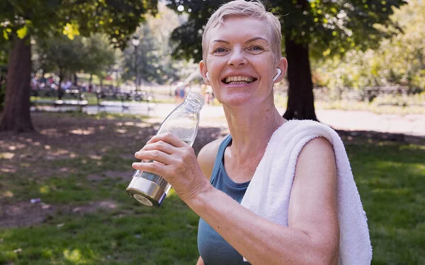 Eine mittelalte Frau mit kurzen blonden Haaren steht nach dem Training lachendlächelnd im Park mit einem über die Schulter geworfenem Handtuch und einer Wasserflasche in der Hand.