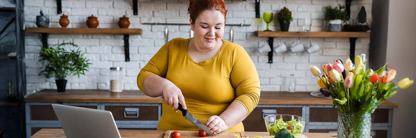 Eine mehrgewichtige Frau mit roten Haaren steht in einer modernen Küche und schneidet auf einem Holzbrett kleine Tomaten. Vor ihr stehen frisches Gemüse wie Brokkoli und Sellerie sowie eine Glasschüssel mit Salat. Neben dem Schneidebrett steht ein geöffneter Laptop. Im Hintergrund sind helle Regale mit Küchenutensilien und Pflanzen zu sehen.