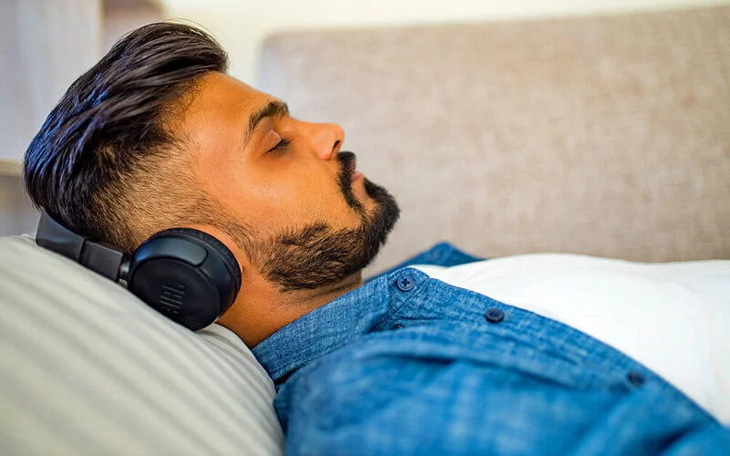 Ein Mann liegt mit erhöhtem Oberkörper und geschlossenen Augen auf der Couch während er über Kopfhörer etwas hört.