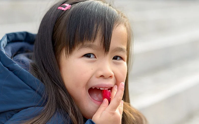 Ein junges Mädchen mit dunklen Haaren, rosa Haarspange und blauer Jacke steckt sich ein rotes Gummibärchen in den Mund.