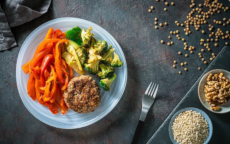 Ein Teller mit Brokkoli, Paprika, Zucchini und einem Fleisch-Patty auf einem Teller. Daneben stehen zwei kleinere Schüsseln mit braunem Reis und Walnüssen.