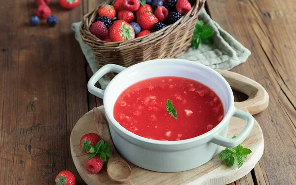 Rote Beeren-Kaltschale auf einem Holztisch, dekoriert und mit Beerenschale dahinter.
