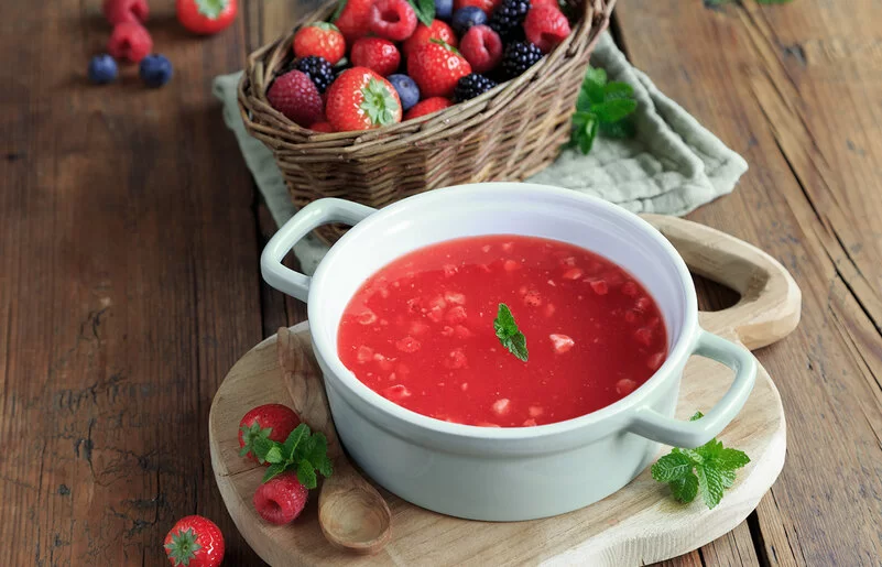 Rote Beeren-Kaltschale auf einem Holztisch, dekoriert und mit Beerenschale dahinter.