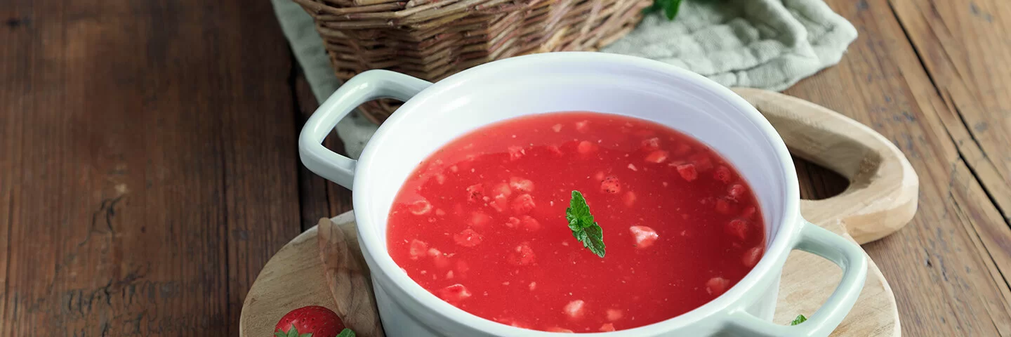Rote Beeren-Kaltschale auf einem Holztisch, dekoriert und mit Beerenschale dahinter.