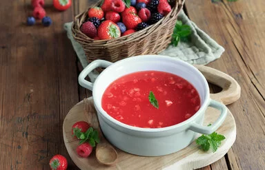 Rote Beeren-Kaltschale auf einem Holztisch, dekoriert und mit Beerenschale dahinter.