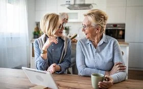 Zwei ältere Frauen sitzen an einem Holztisch in einer modernen, hellen Küche. Beide tragen helle Blusen und haben kurze, helle Haare. Eine Frau hält eine Tasse in der Hand, während die andere mit einer Hand gestikuliert. Auf dem Tisch steht ein Tablet mit weißer Hülle. Im Hintergrund ist ein älterer Mann unscharf zu erkennen.
