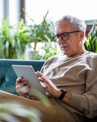Ein älterer Mann sitzt auf der Couch in seinem Wohnzimmer und blickt interessiert auf ein Tablet, das er in den Händen hält.