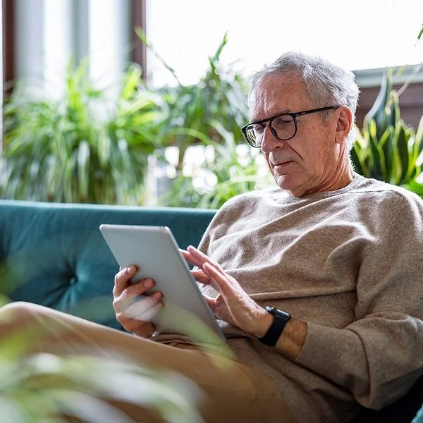 Ein älterer Mann sitzt auf der Couch in seinem Wohnzimmer und blickt interessiert auf ein Tablet, das er in den Händen hält.