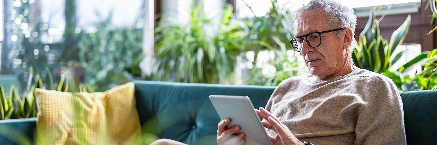 Ein älterer Mann sitzt auf der Couch in seinem Wohnzimmer und blickt interessiert auf ein Tablet, das er in den Händen hält.