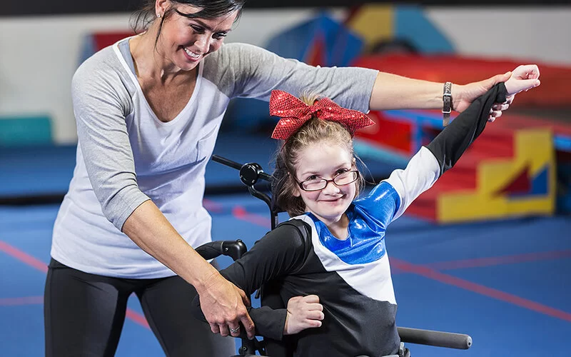 Ein Mädchen im Rollstuhl macht eine Cheerleading-Pose, unterstützt durch eine Trainerin.