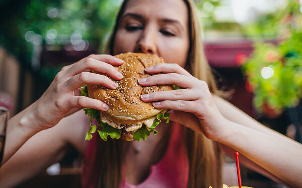 Eine junge blonde Frau hält einen fetten Burger in den Händen und beißt hinein.