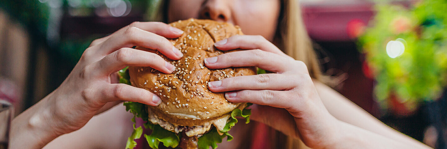 Eine junge blonde Frau hält einen fetten Burger in den Händen und beißt hinein.