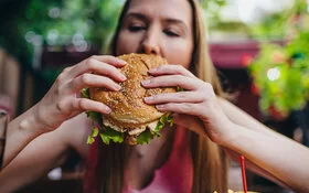 Eine junge blonde Frau hält einen fetten Burger in den Händen und beißt hinein.