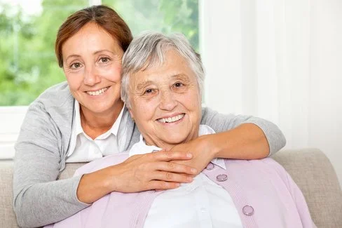 Eine Frau mittleren Alters umarmt ihre pflegebedürftige Mutter während beide in die Kamera lächeln.