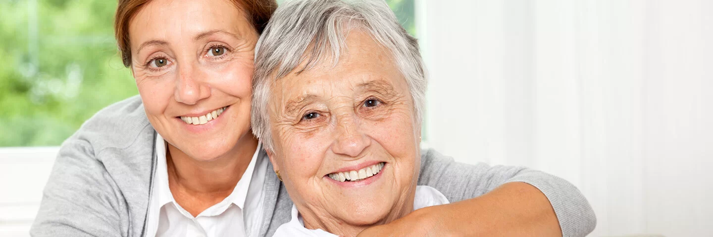 Eine Frau mittleren Alters umarmt ihre pflegebedürftige Mutter während beide in die Kamera lächeln.