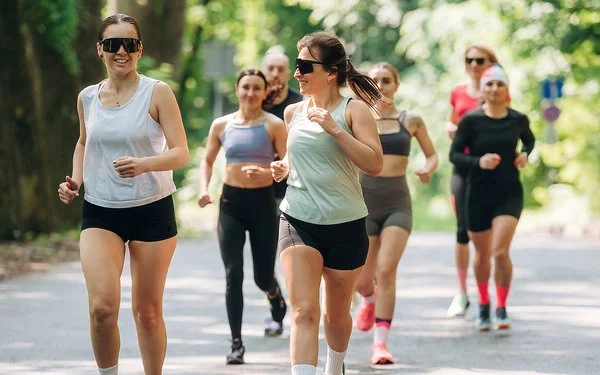 Läuferinnen und Läufer eines Running Clubs joggen gemeinsam auf einer sonnigen Straße durch einen grünen Wald.