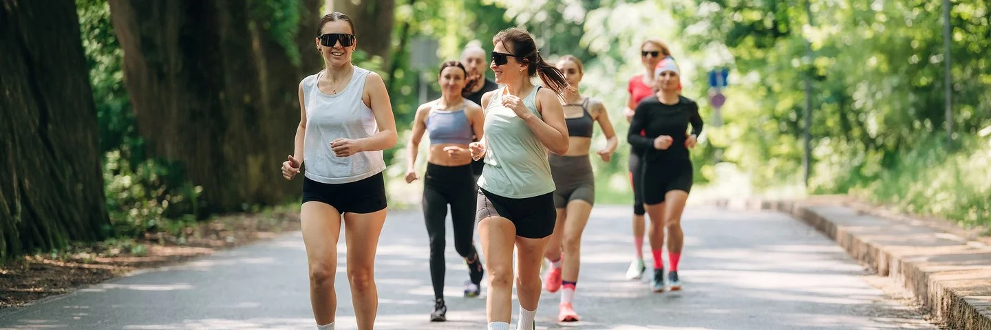 Läuferinnen und Läufer eines Running Clubs joggen gemeinsam auf einer sonnigen Straße durch einen grünen Wald.