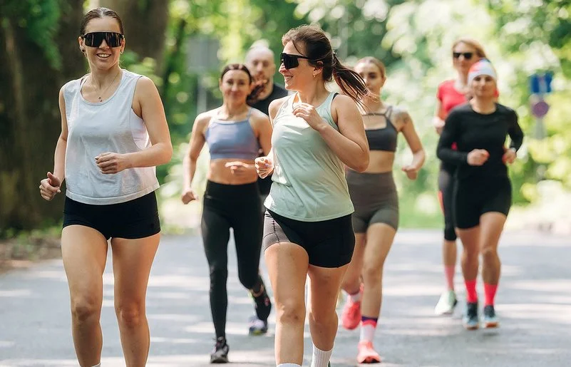 Läuferinnen und Läufer eines Running Clubs joggen gemeinsam auf einer sonnigen Straße durch einen grünen Wald.
