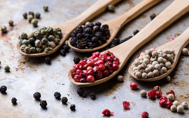 Auf einer Metalloberfläche liegen vier Holzlöffel. Jeder Löffel ist mit einer anderen Pfeffersorte gefüllt: grüne, schwarze, rote und weiße Pfefferkörner.