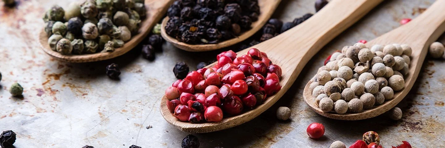 Auf einer Metalloberfläche liegen vier Holzlöffel. Jeder Löffel ist mit einer anderen Pfeffersorte gefüllt: grüne, schwarze, rote und weiße Pfefferkörner.