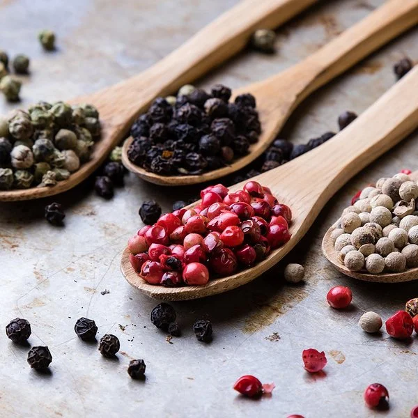 Auf einer Metalloberfläche liegen vier Holzlöffel. Jeder Löffel ist mit einer anderen Pfeffersorte gefüllt: grüne, schwarze, rote und weiße Pfefferkörner.